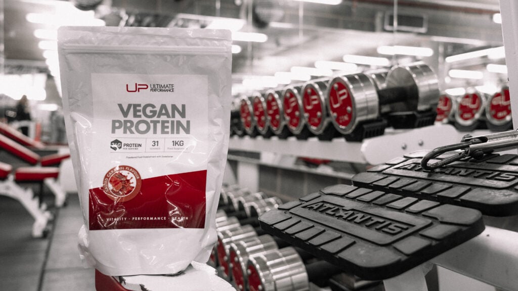 A pouch of Ultimate Performance vegan protein powder on a gym backdrop with weights