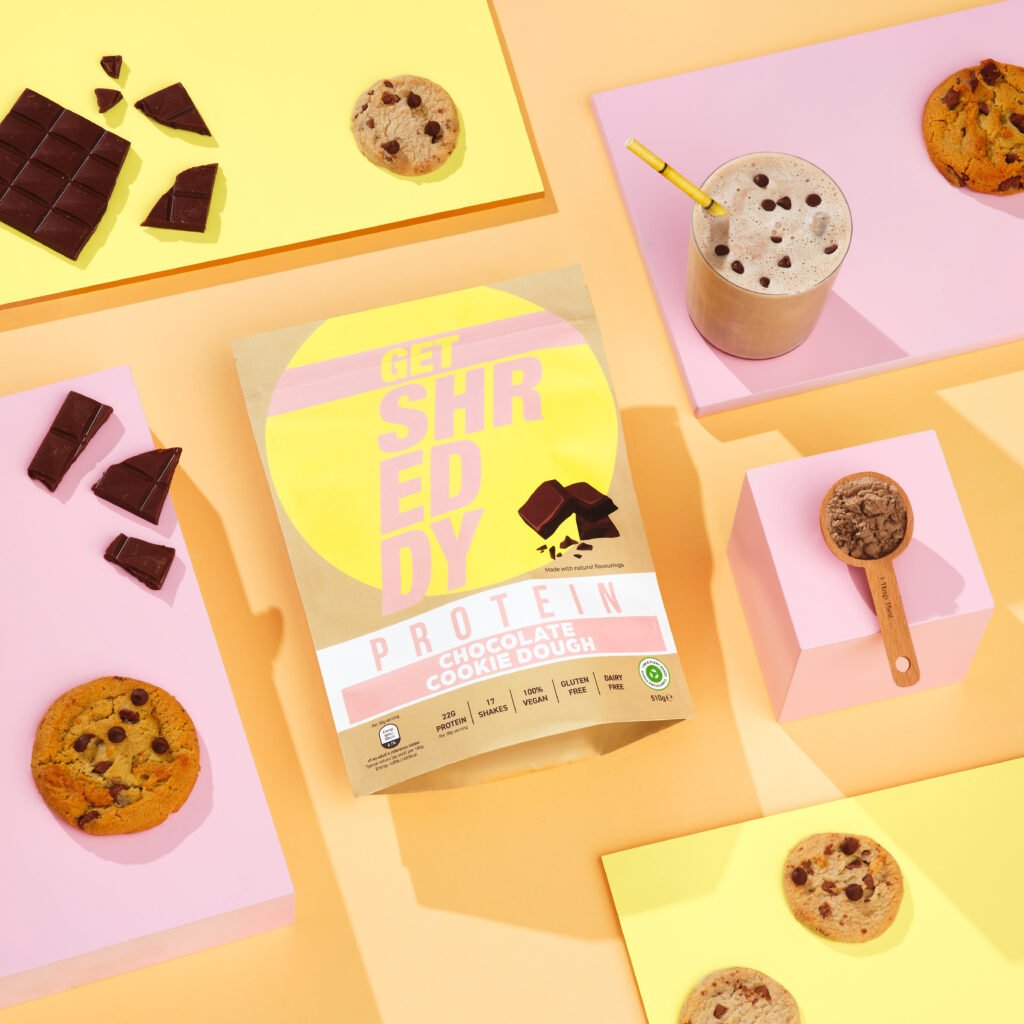 A packet of Shreddy vegan protein powder on a pastel background with cookies, chocolate and a prepared drink in shot