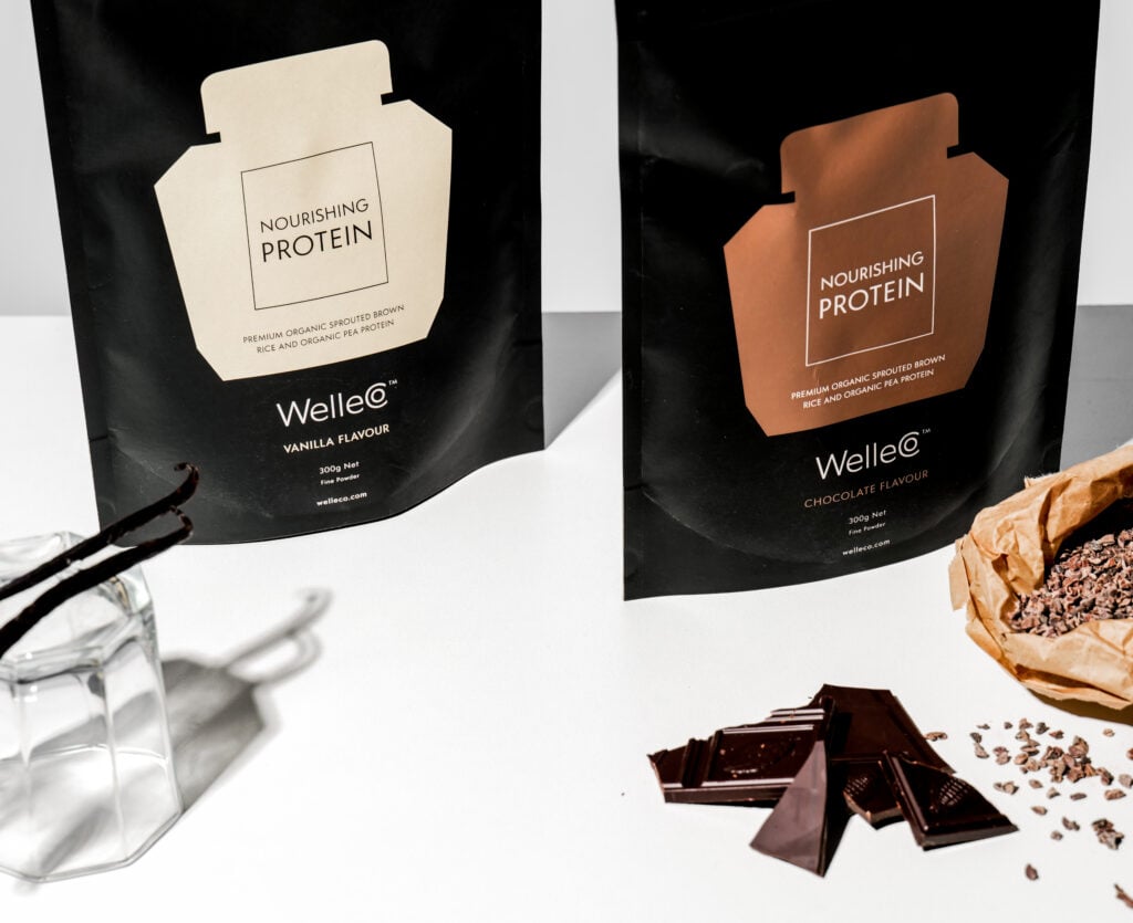 WelleCo vegan protein powder packets on a white table with chocolate and vanilla pods in shot