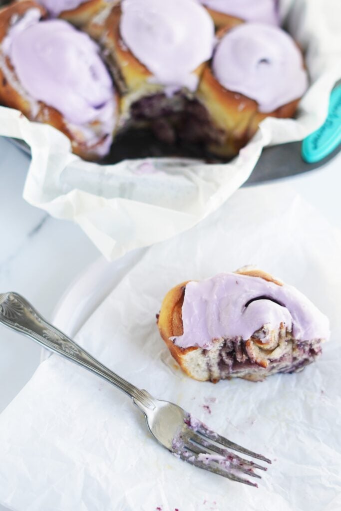 a baking tin full of freshly iced vegan blueberry cinnamon rolls with one in the foreground, partially eaten