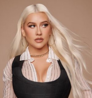 Christina Aguilera standing in front of a neutral background wearing a white shirt and black corset top with long straight blonde hair