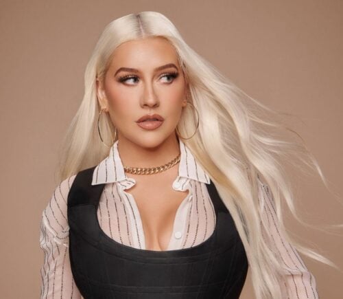 Christina Aguilera standing in front of a neutral background wearing a white shirt and black corset top with long straight blonde hair