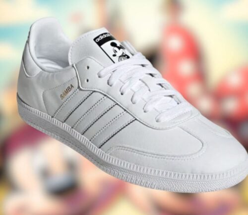All-white vegan Adidas x Disney Samba sneakers with Mickey Mouse motifs, with a blurred image of Mickey and Minnie in the background