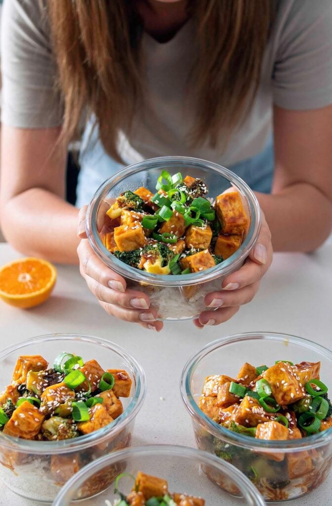 bowls of freshly prepared orange tofu and broccoli, one being held by the recipe developer from Plant Baes
