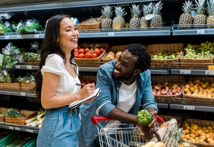 A couple laughing in the fruit and vegetable aisle of a supermarket, pushing a shopping cart