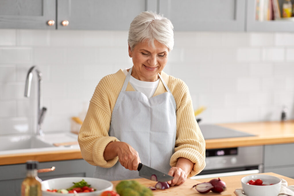An older woman chopping up a red onion while making a salad in a well-lit kitchen