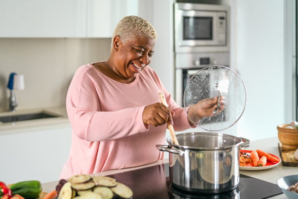 An older woman smiling and cooking vegetables in a large pot in a bright kitchen