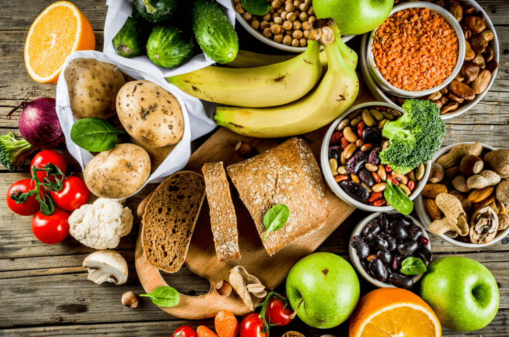 Healthy plant-based food including fruit, vegetables, legumes, nuts, and bread spread out on a wooden table