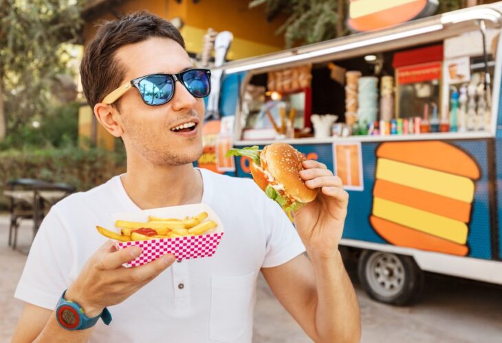 A man eating a meat burger in front of a food truck and wearing polarized sunglasses