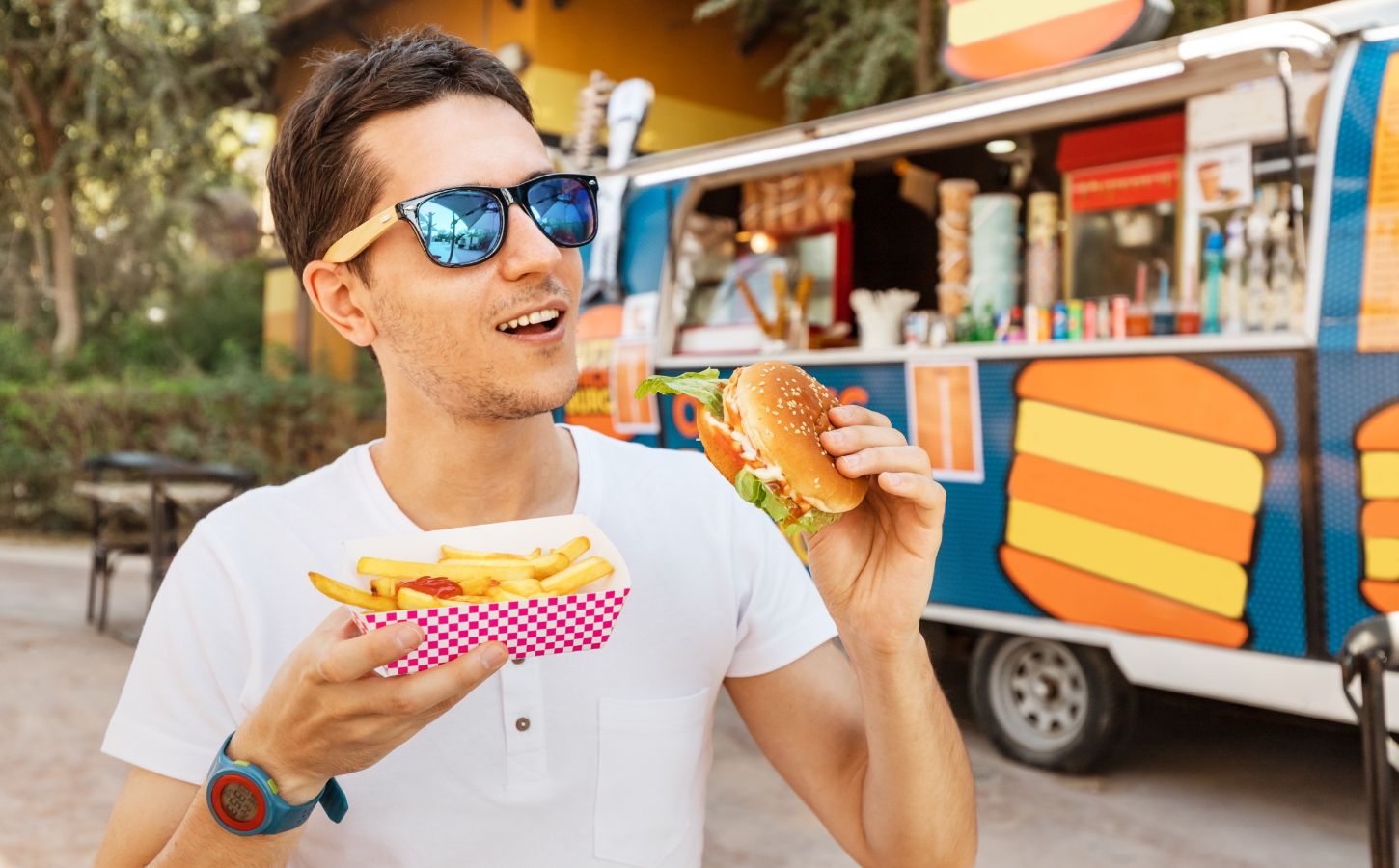 A man eating a meat burger in front of a food truck and wearing polarized sunglasses