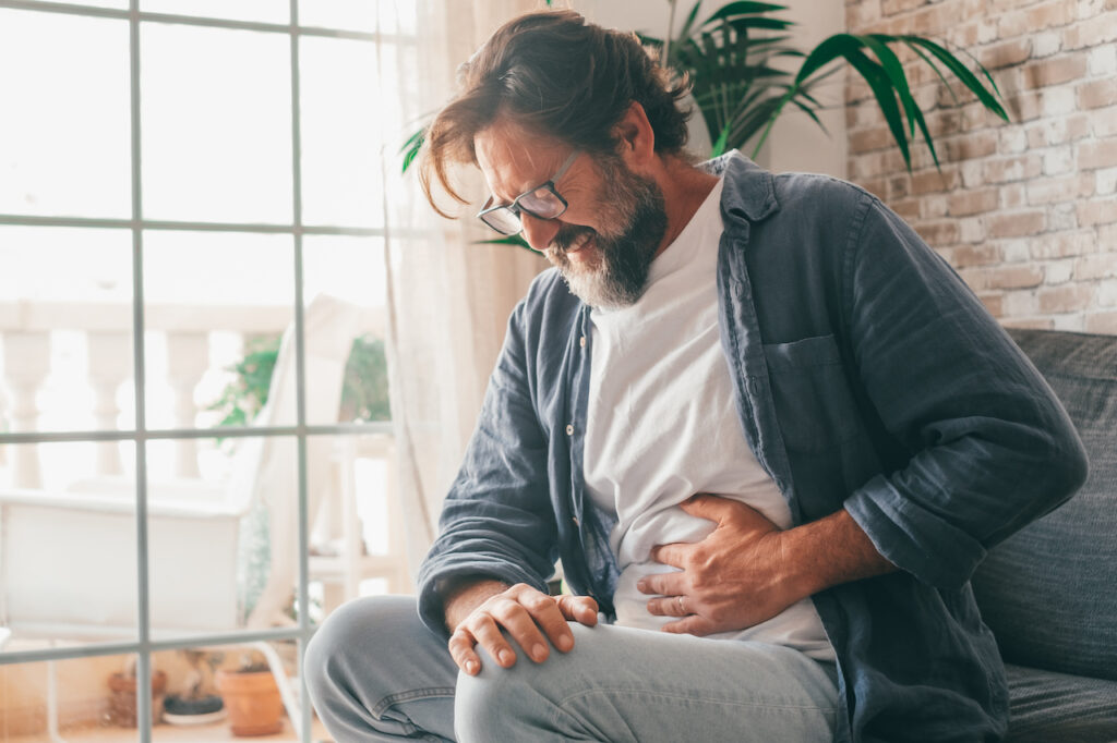 A middle-aged man looks in pain and clutches his stomach while sat on a sofa