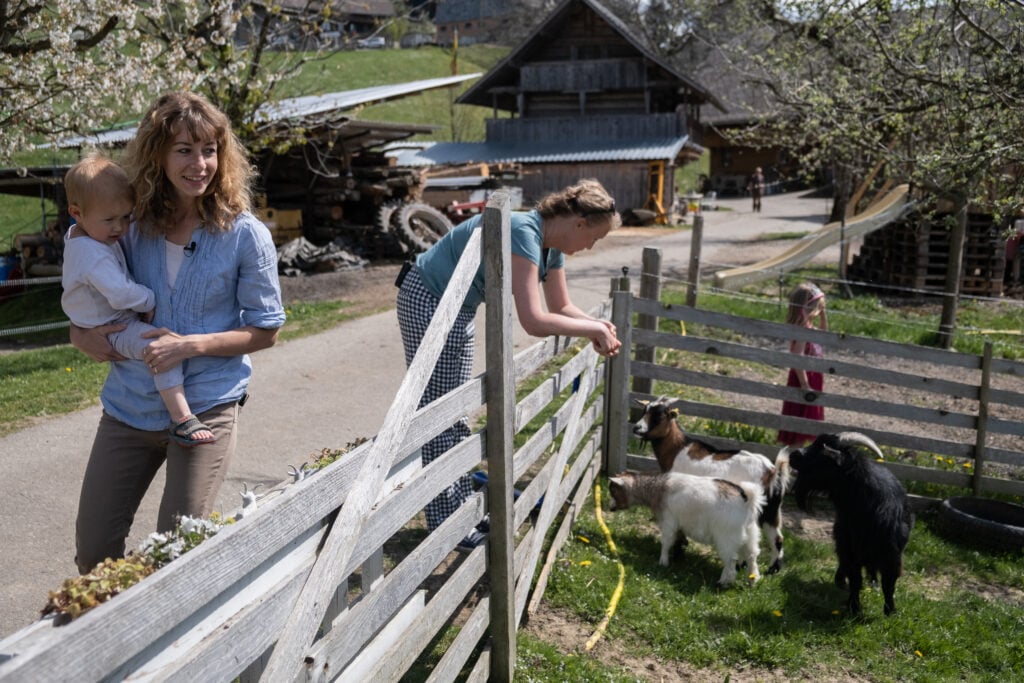 A former beef farm owner Selina holding a baby next to an animal pen, while Sarah Heiligtag stands next to her feeding the animals