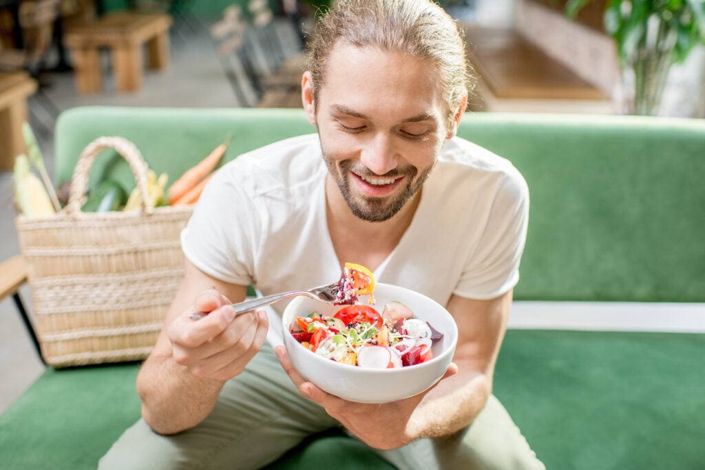 Handsome man eating healthy salad sitting indoors on the green sofa with bag full of vegetables on the background. Healthy eating concept