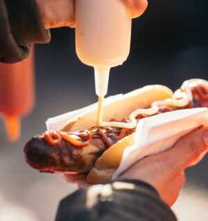 A person's hand squeezing sauce onto a meat hot dog, which can raise stroke risk