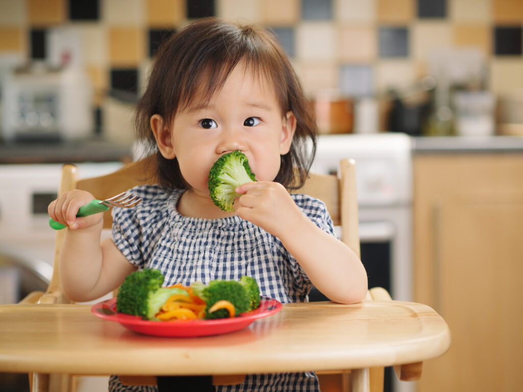 A young child eating broccoli and other vegetables while sat in a high chair