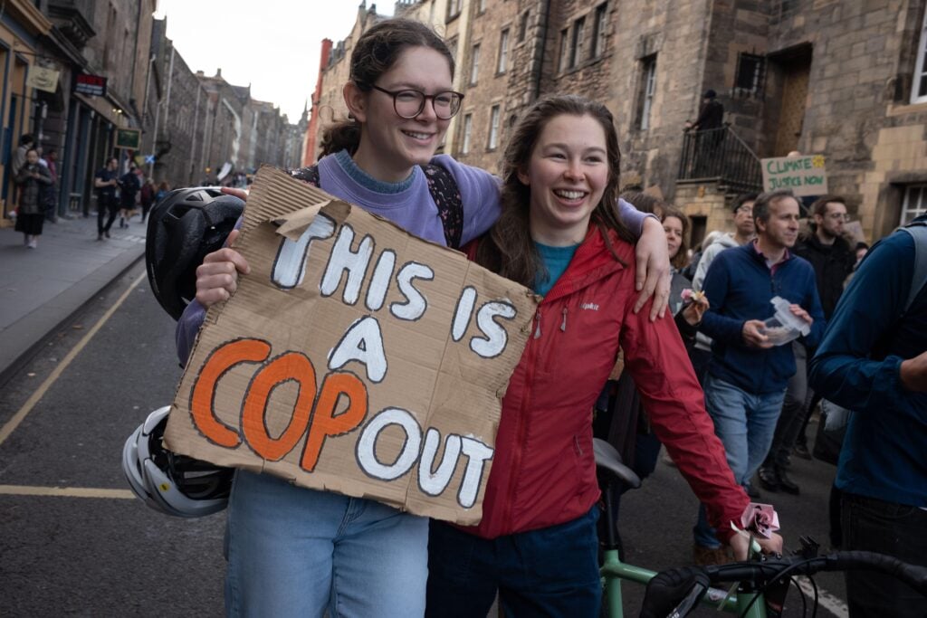Two young people protest outside COP with a sign saying "This is a COP out"