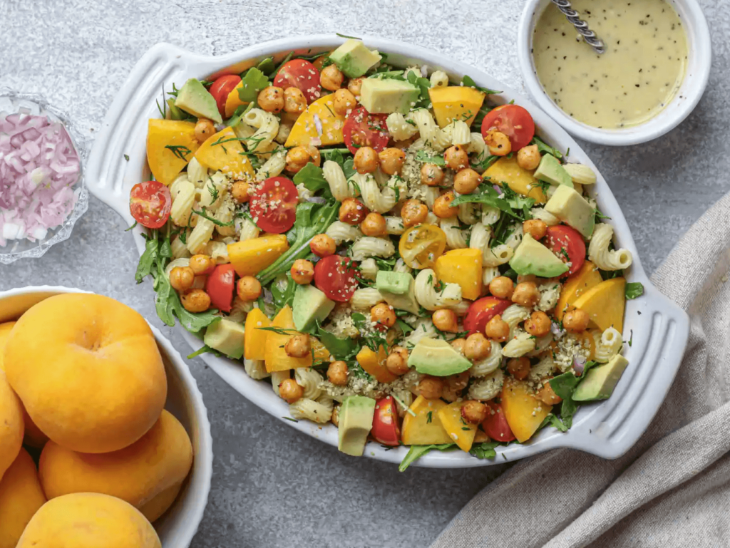 A dish of pasta salad made from a vegan recipe, featuring peach, rocket, tomato, and chickpeas