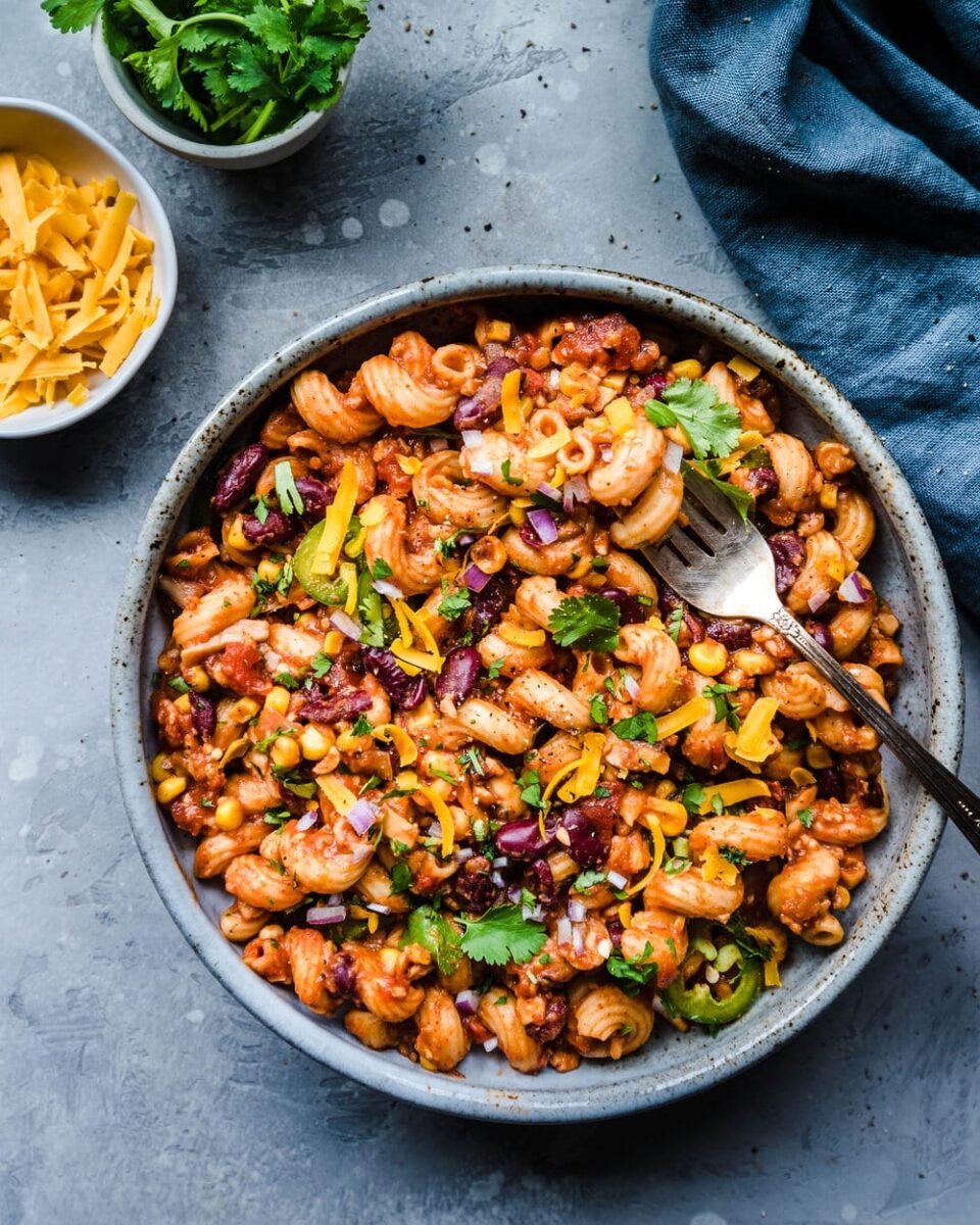 A vegan chili mac in a bowl next to dairy-free cheese and cilantro