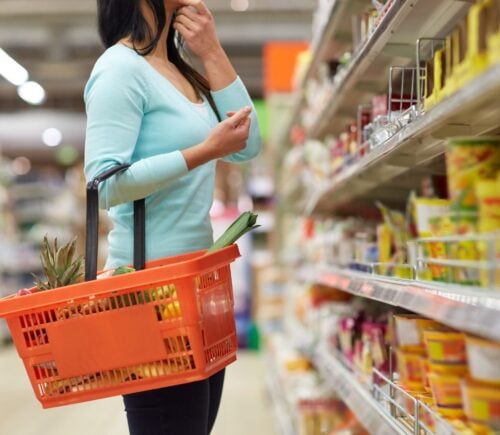 Photo shows a woman shopping in a supermarket looking at the shelves and holding an orange basket.
