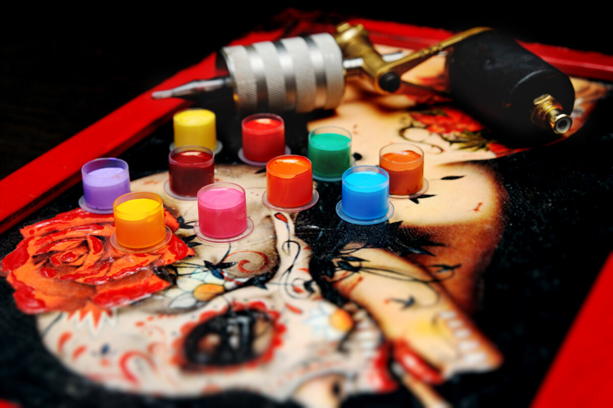 Photo shows a selection of brightly colored tattoo inks next to a tattoo machine on a surface decorated with a Day of the Dead-style woman.