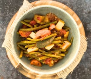A bowl of green bean stew on top of a wooden board on a dark countertop