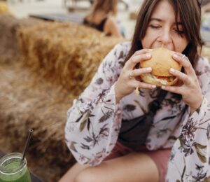 Photo shows a young woman sat on a hay bale and taking a bite out of a burger