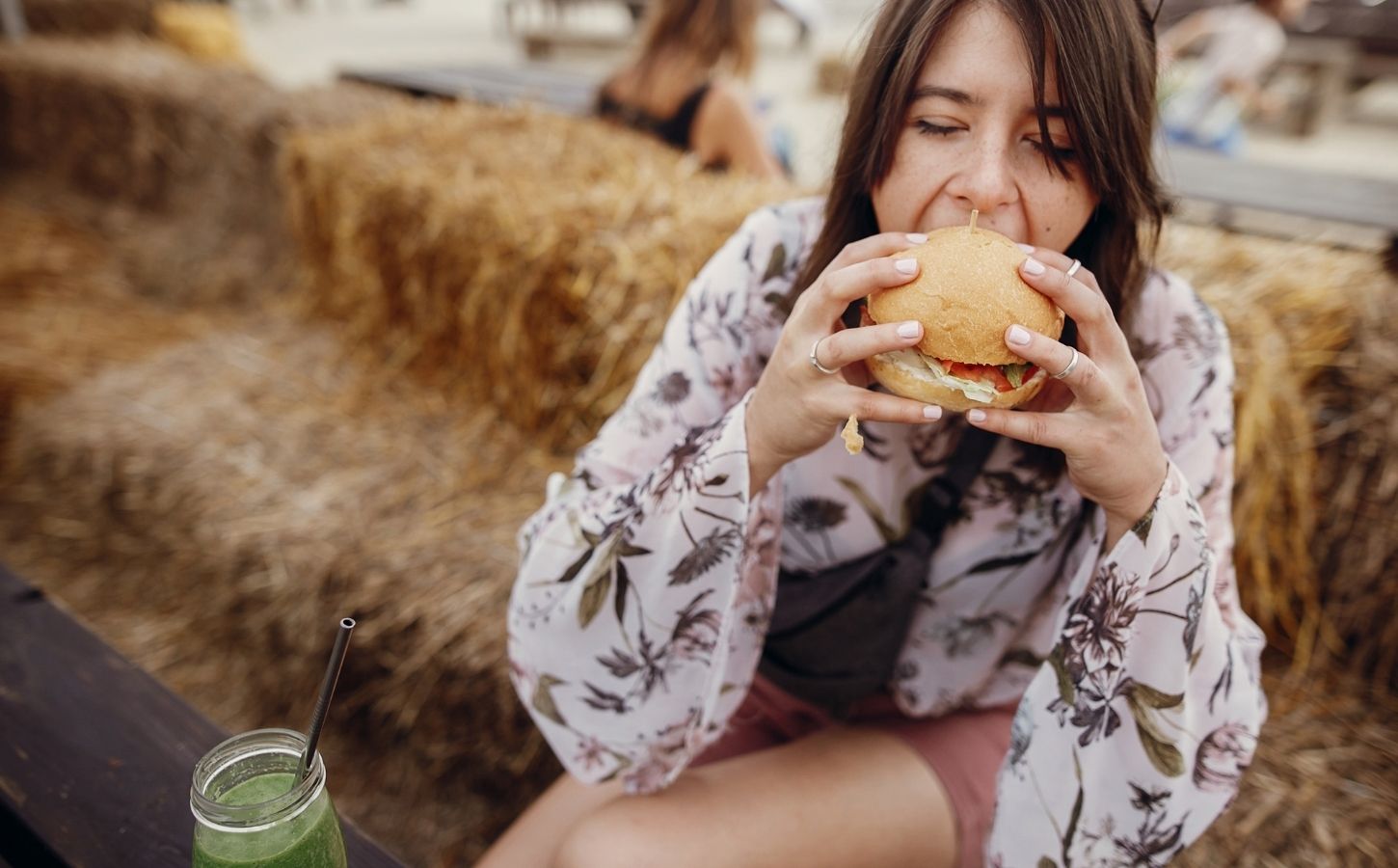 Photo shows a young woman sat on a hay bale and taking a bite out of a burger