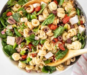 A Greek pasta salad made with dairy-free feta cheese, filled with tomatoes, olives, and spinach