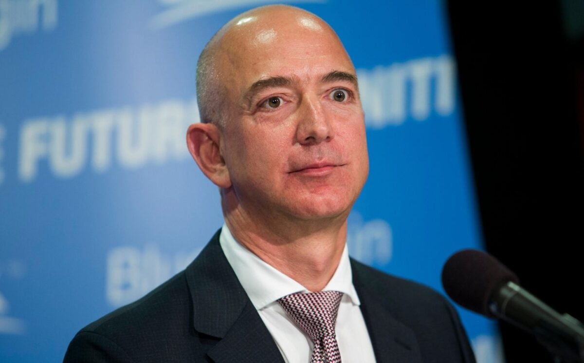 Photo shows a photo of Amazon founder Jeff Bezos speaking into a microphone against a blue backdrop