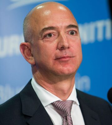 Photo shows a photo of Amazon founder Jeff Bezos speaking into a microphone against a blue backdrop