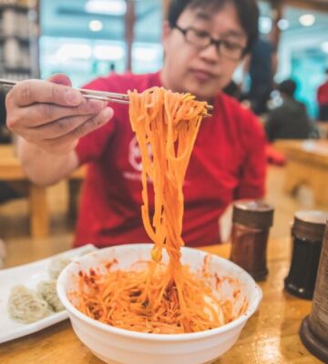 Photo shows a man holding up noodles over a bowl using chopsticks