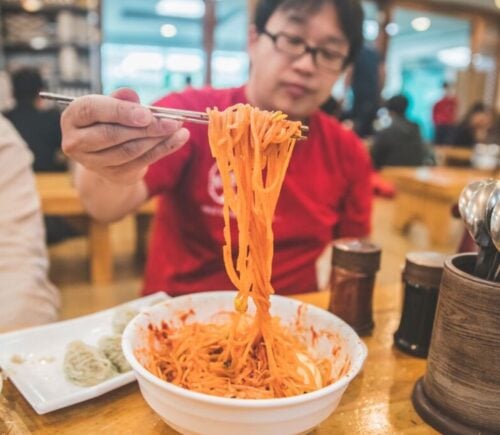 Photo shows a man holding up noodles over a bowl using chopsticks