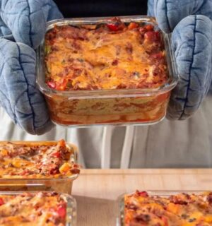 Photo shows someone holding a large tray of vegan lasagna in pale blue oven gloves, with several more trays already on the table