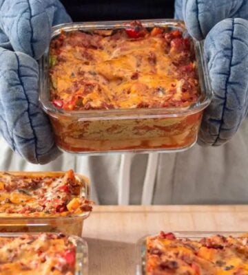 Photo shows someone holding a large tray of vegan lasagna in pale blue oven gloves, with several more trays already on the table