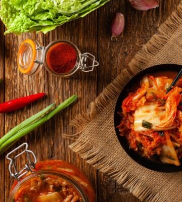 Photo shows a bowl of kimchi on a wooden tabletop surrounded by common kimchi-making ingredients like scallions, chili, and ginger