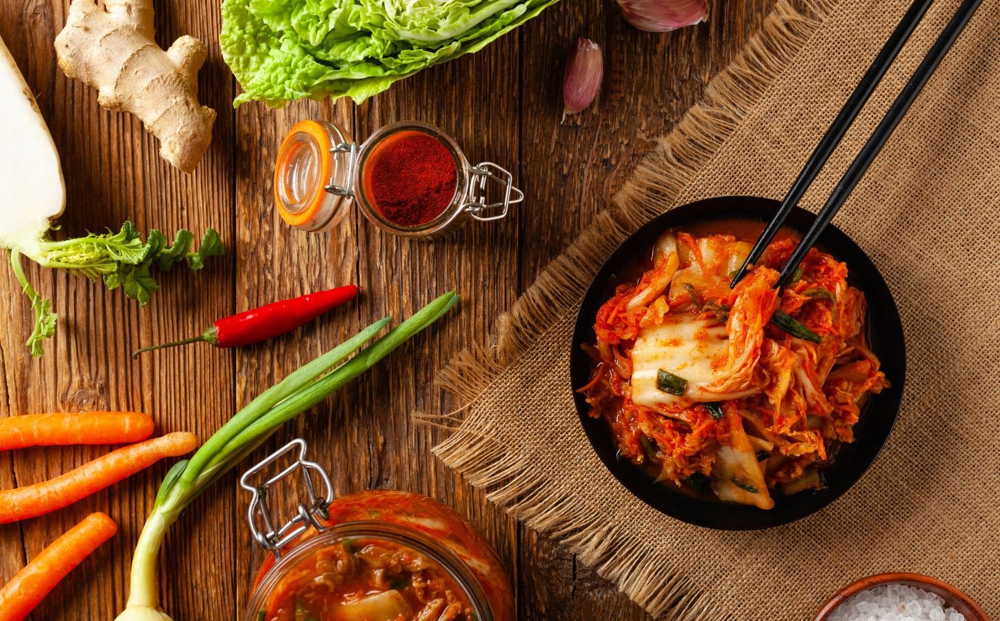 Photo shows a bowl of kimchi on a wooden tabletop surrounded by common kimchi-making ingredients like scallions, chili, and ginger