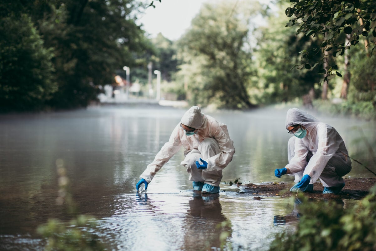 Photo shows two scientists in protecting suits standing in a river and taking water samples