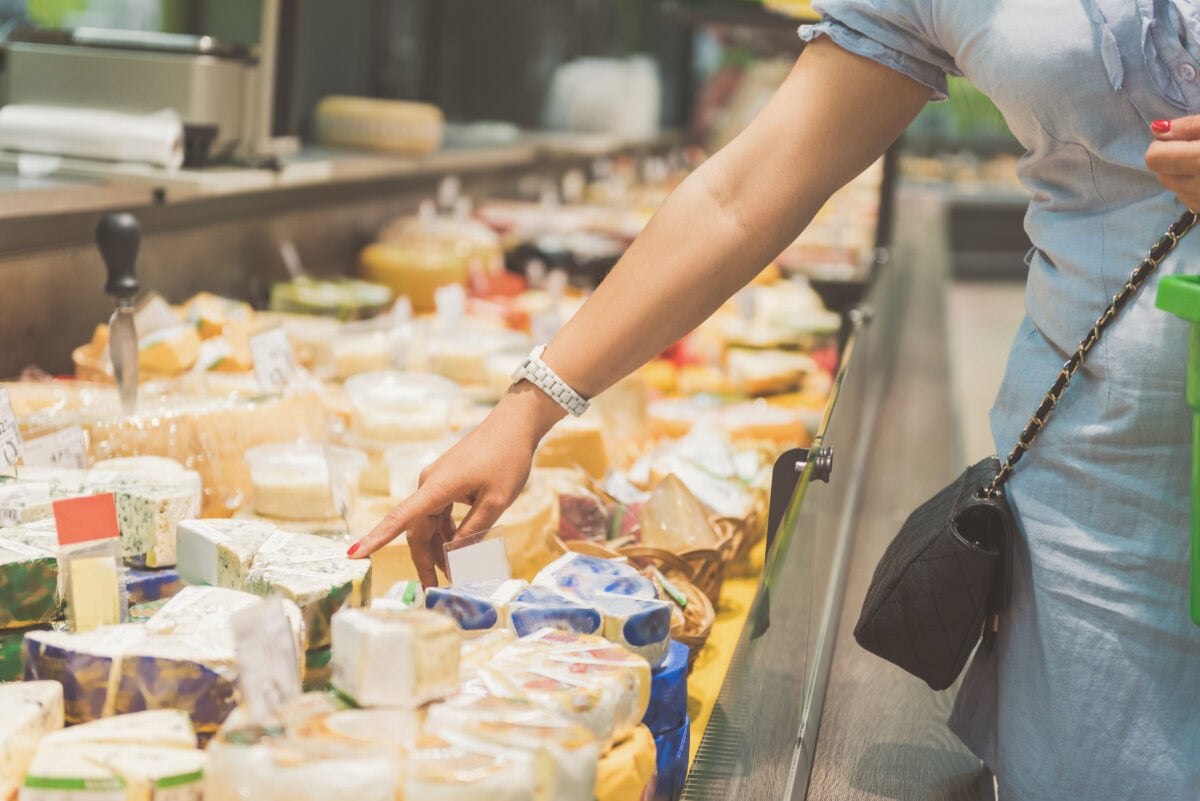 Photo shows a woman's arm and part of her torso as she gestures to a particular piece of cheese at a supermarket dairy counter