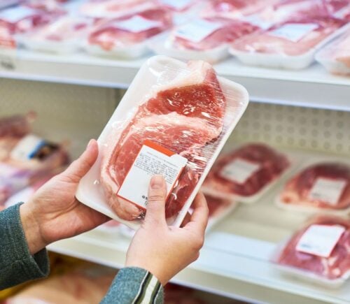 Photo shows someone holding up a packet of pork in front of a supermarket shelf stocked with the same