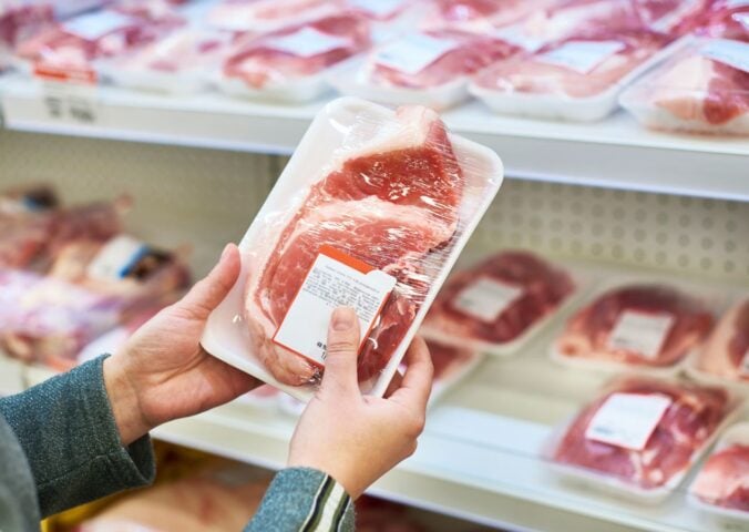 Photo shows someone holding up a packet of pork in front of a supermarket shelf stocked with the same