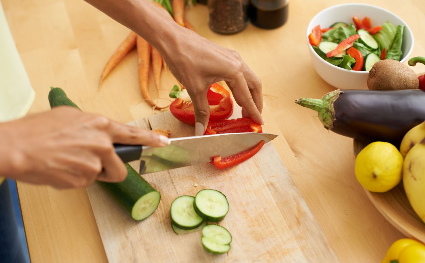 Photo shows someone's hands as they prepare fresh vegetables and produce with a knife on a wooden chopping board