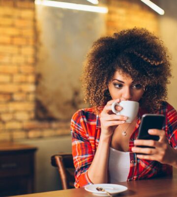Photo shows a woman drinking coffee at a wooden dining table and holding her phone up in one hand