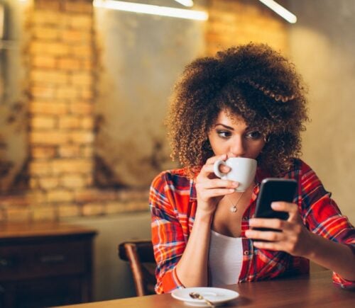 Photo shows a woman drinking coffee at a wooden dining table and holding her phone up in one hand