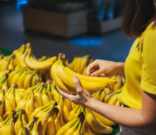 Photo shows a woman in a yellow shirt picking up a bunch of bananas from a large display case full of the same in a grocery store
