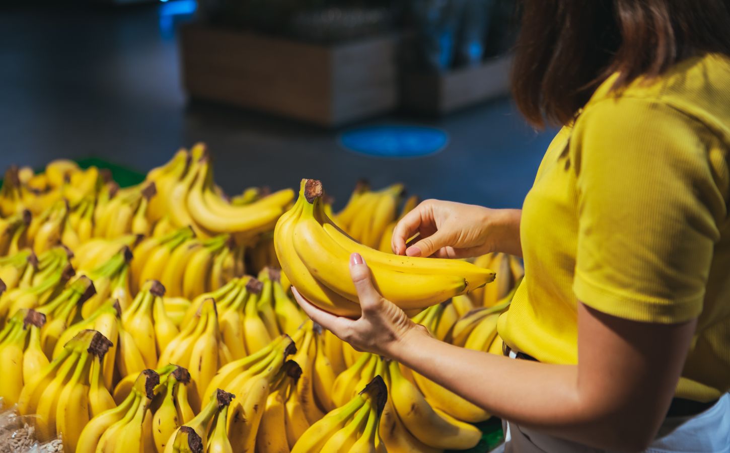 Photo shows a woman in a yellow shirt picking up a bunch of bananas from a large display case full of the same in a grocery store