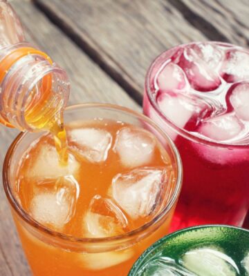 Photo shows a bottle of bright orange soda being poured into a glass with ice, next to three others which are red, green, and black