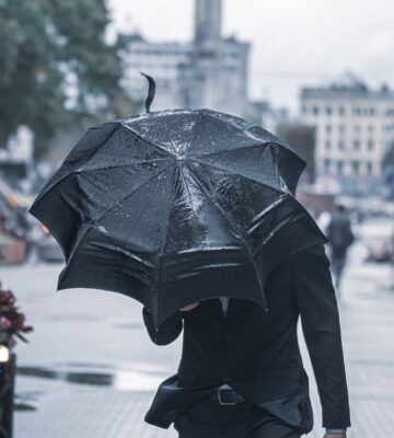 Photo shows someone in a suit walking with a black umbrella held low against heavy rain
