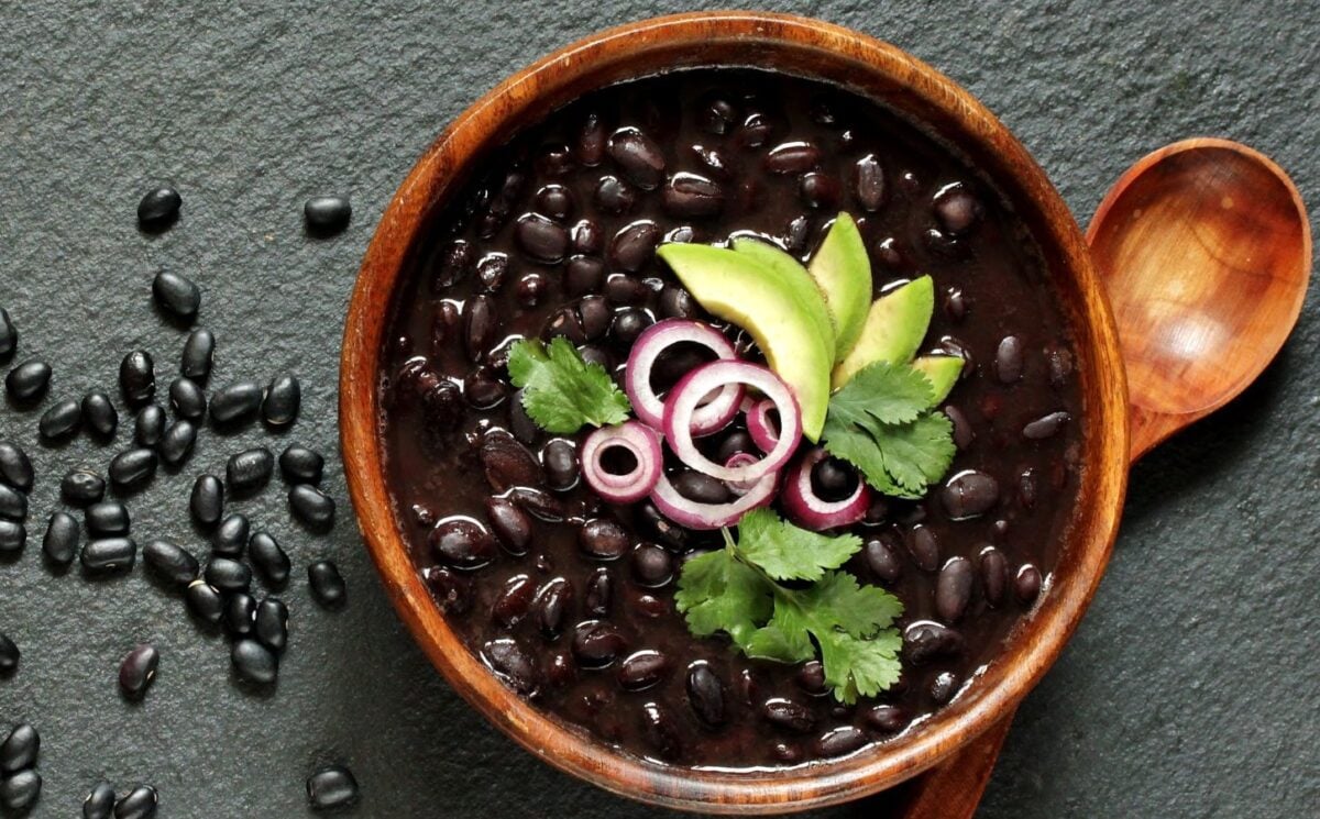Photo shows a wooden bowl of thick black bean stew or soup, decorated with red onion, corander, and avocado