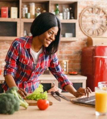 A woman in a brightly lit kitchen chopping up salad next to a laptop