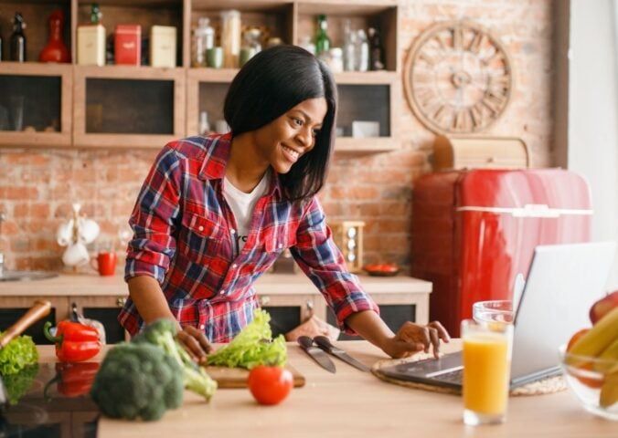 A woman in a brightly lit kitchen chopping up salad next to a laptop